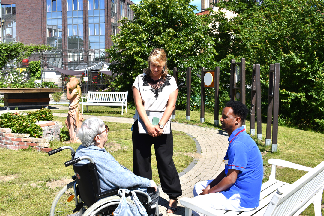 Die Bildungsbeauftragte, ein Auszubildender und eine Bewohnerin unterhalten sich auf dem Hof in der Sonne.